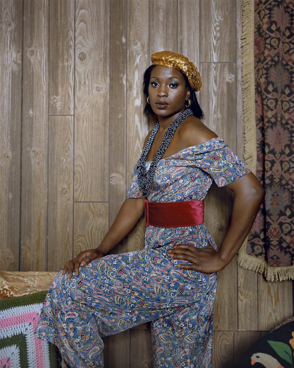 Person wearing a patterned dress, red belt, and gold hat, posing with one hand on hip against a wood-paneled wall.