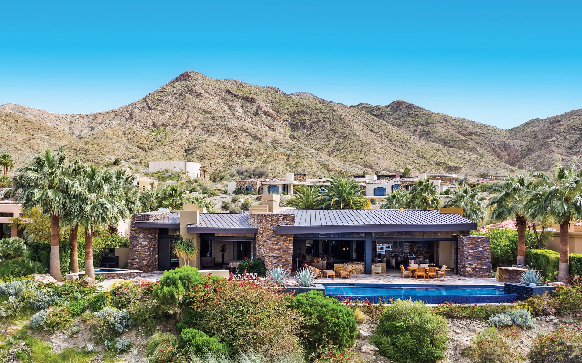 Luxury modern villa with large pool set against a backdrop of desert mountains and palm trees on a sunny day.