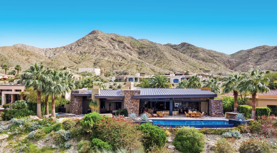 Luxury desert home with pool, surrounded by palm trees and mountain landscape in the background.