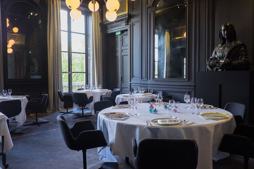 Elegant dining room with round tables, white tablecloths, black chairs, large windows, and a decorative statue in dim lighting.