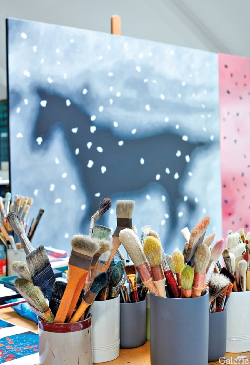 Paintbrushes in containers on a table with a partially painted canvas of a horse in the background.