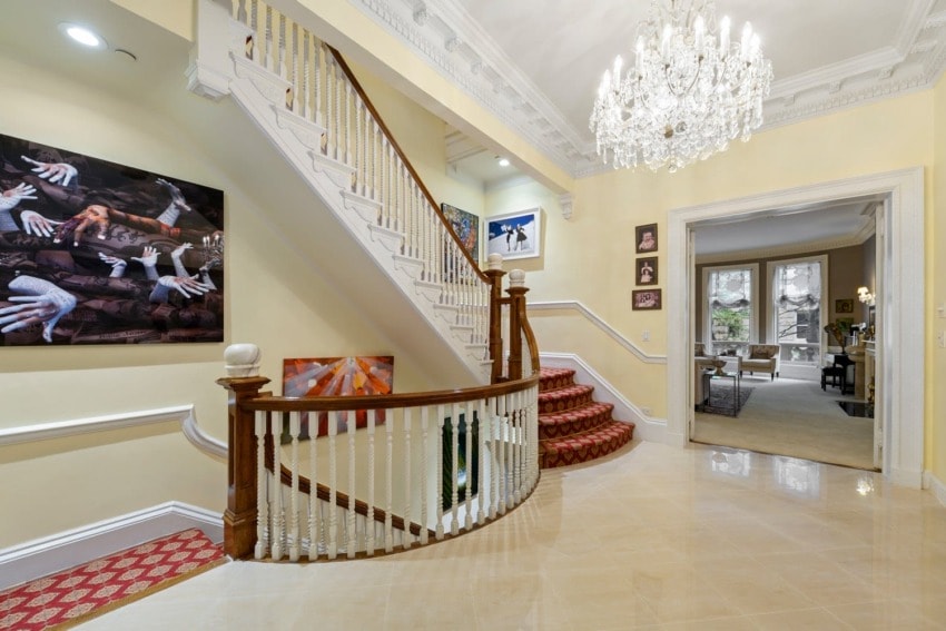 Elegant foyer with a grand staircase, chandelier, and artwork on walls, leading to a spacious living area with large windows.