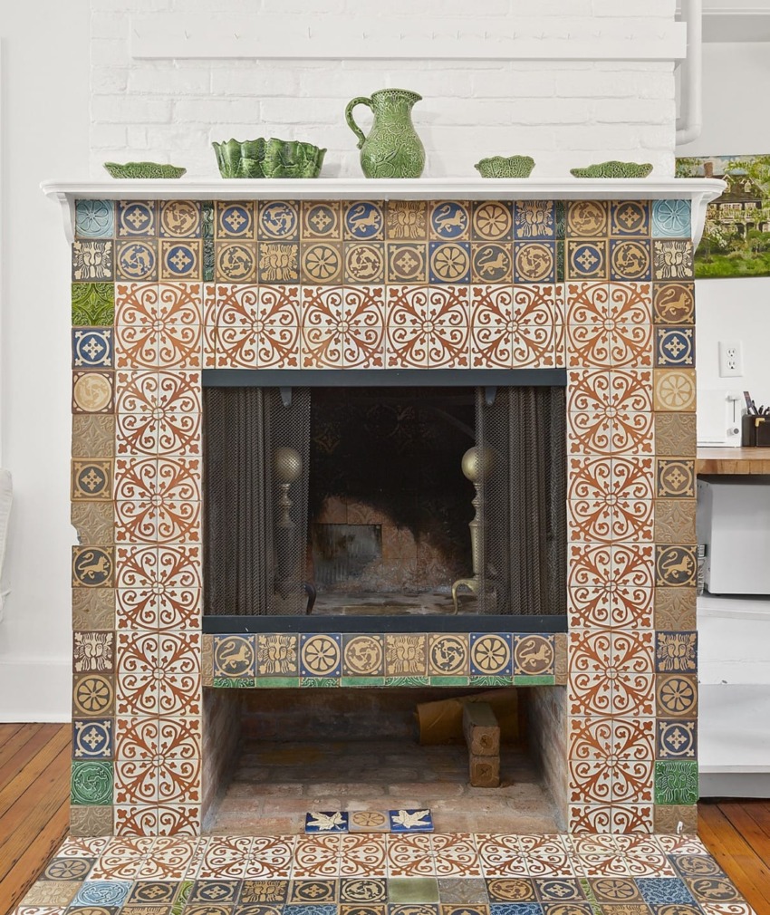 Colorful tiled fireplace with intricate patterns and a green jug on the mantel against a white brick wall.
