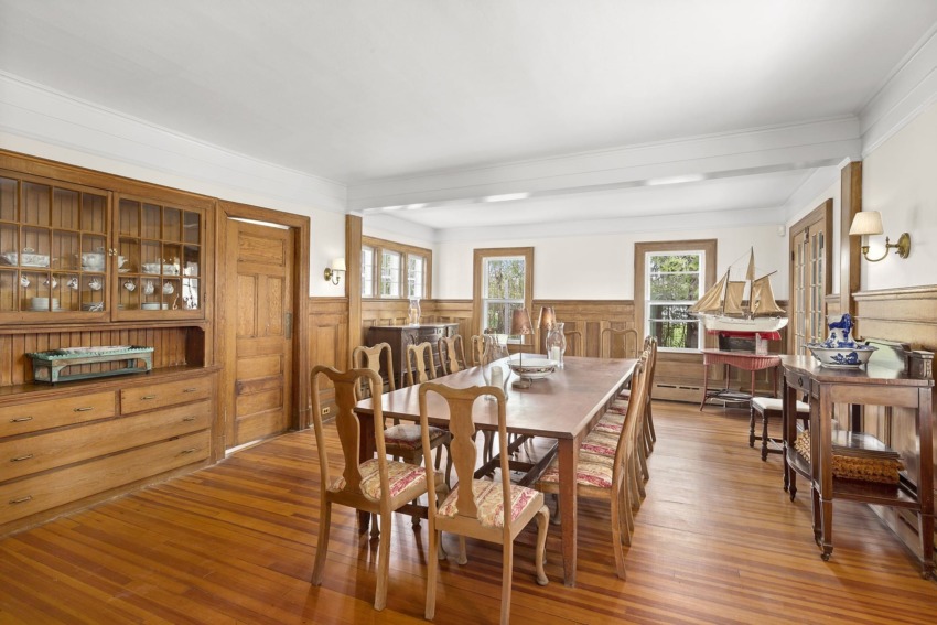 Spacious dining room with long wooden table, chairs, wooden cabinets, sailboat model, and large windows.