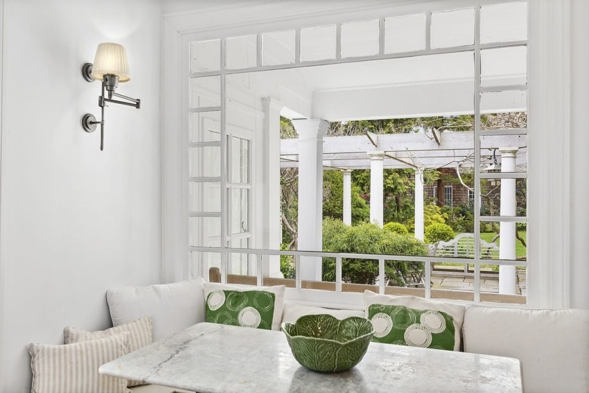 Cozy dining area with white bench seating and green cushions, overlooking a lush garden through a large window.