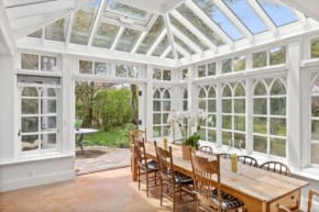 Bright sunroom with large windows, long wooden dining table, chairs, and potted orchids, leading to a garden patio.