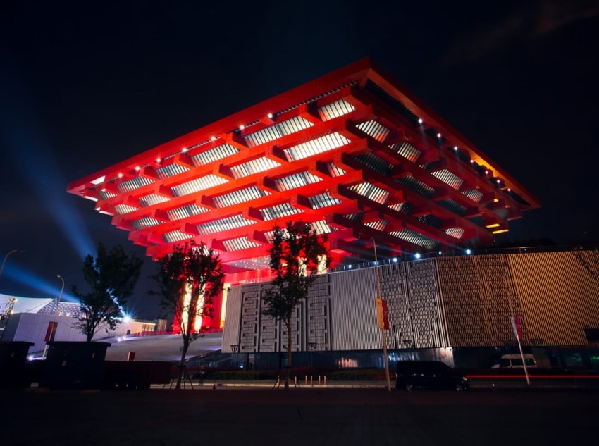 Night view of a brightly illuminated modern architectural building with red accents and trees in the foreground.