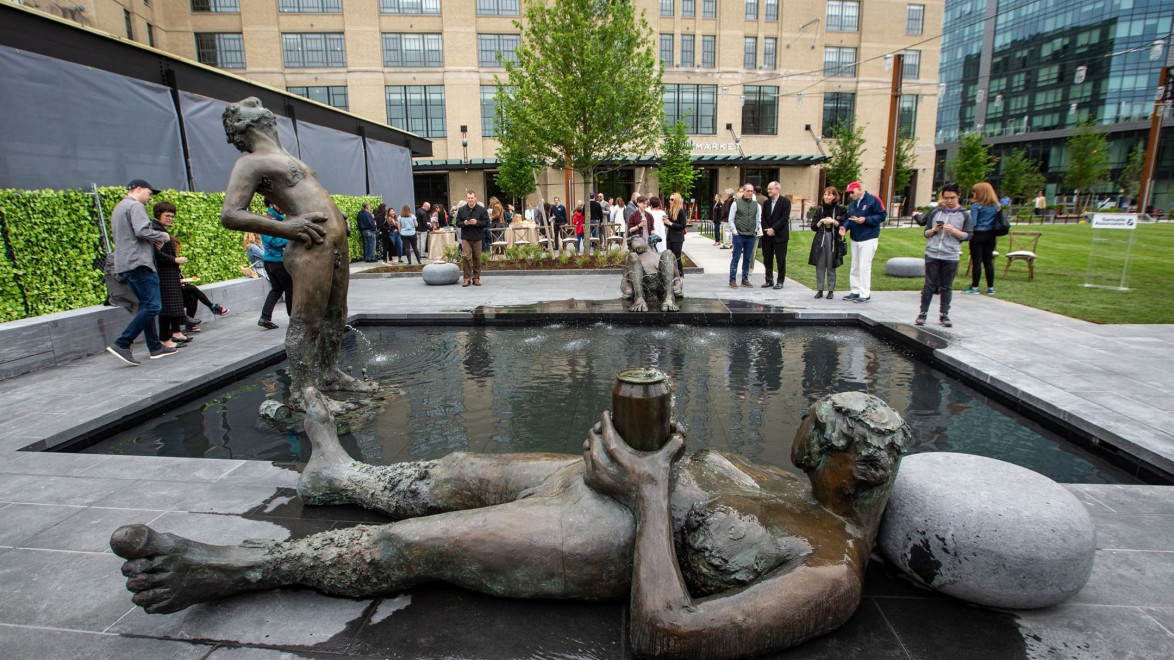 People gather around a fountain with several unique sculptures in a modern urban park setting.