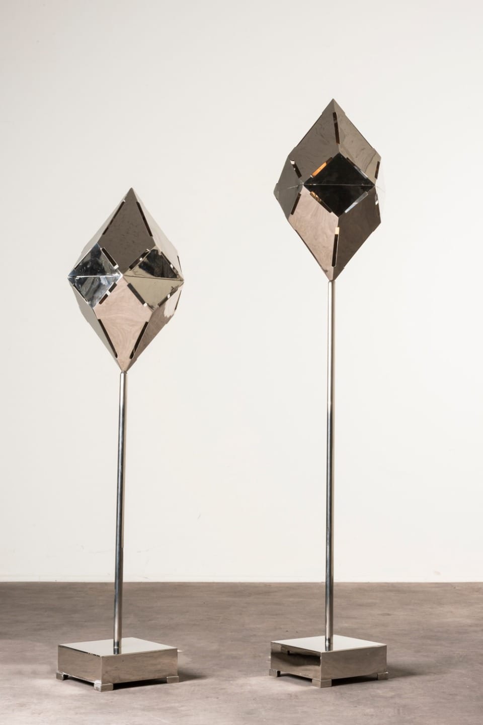 Two modern geometric sculptures with reflective surfaces on stands against a plain background.