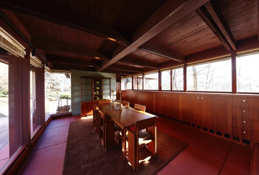 Interior of a mid-century modern dining room with wooden walls, ceiling beams, and large windows overlooking a garden.