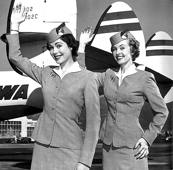 Flight attendants in vintage uniforms smiling and waving in front of an airplane, capturing mid-20th century aviation style.