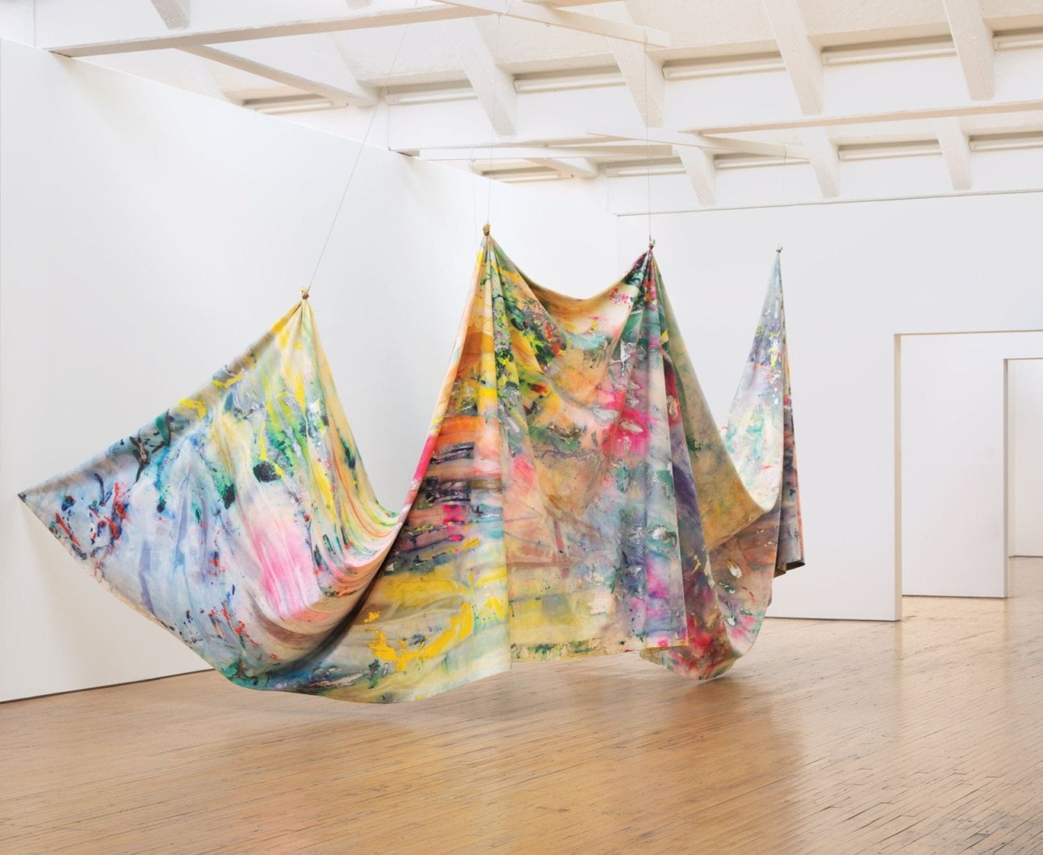 Colorful abstract fabric art installation hanging in a bright gallery space with wooden floor and white walls.
