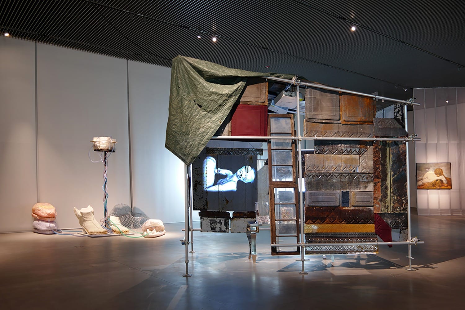 Contemporary art installation with various materials, including tarpaulin, metal structures, wires, and mixed media objects.