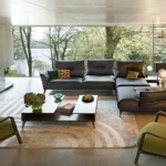 Modern living room with green chairs, grey sectional sofa, fireplace, large windows with a forest view, and ambient lighting.