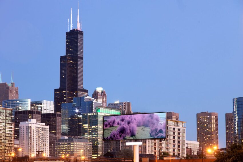 Chicago skyline at dusk with Willis Tower and city buildings, foreground digital billboard displaying colorful image.