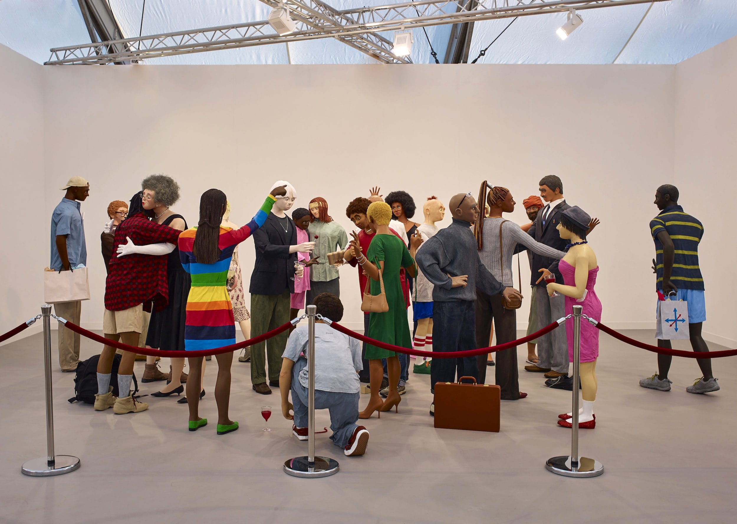 Lifelike sculptures of diverse people at an art exhibit, gathered in a realistic, everyday scenario behind red ropes.