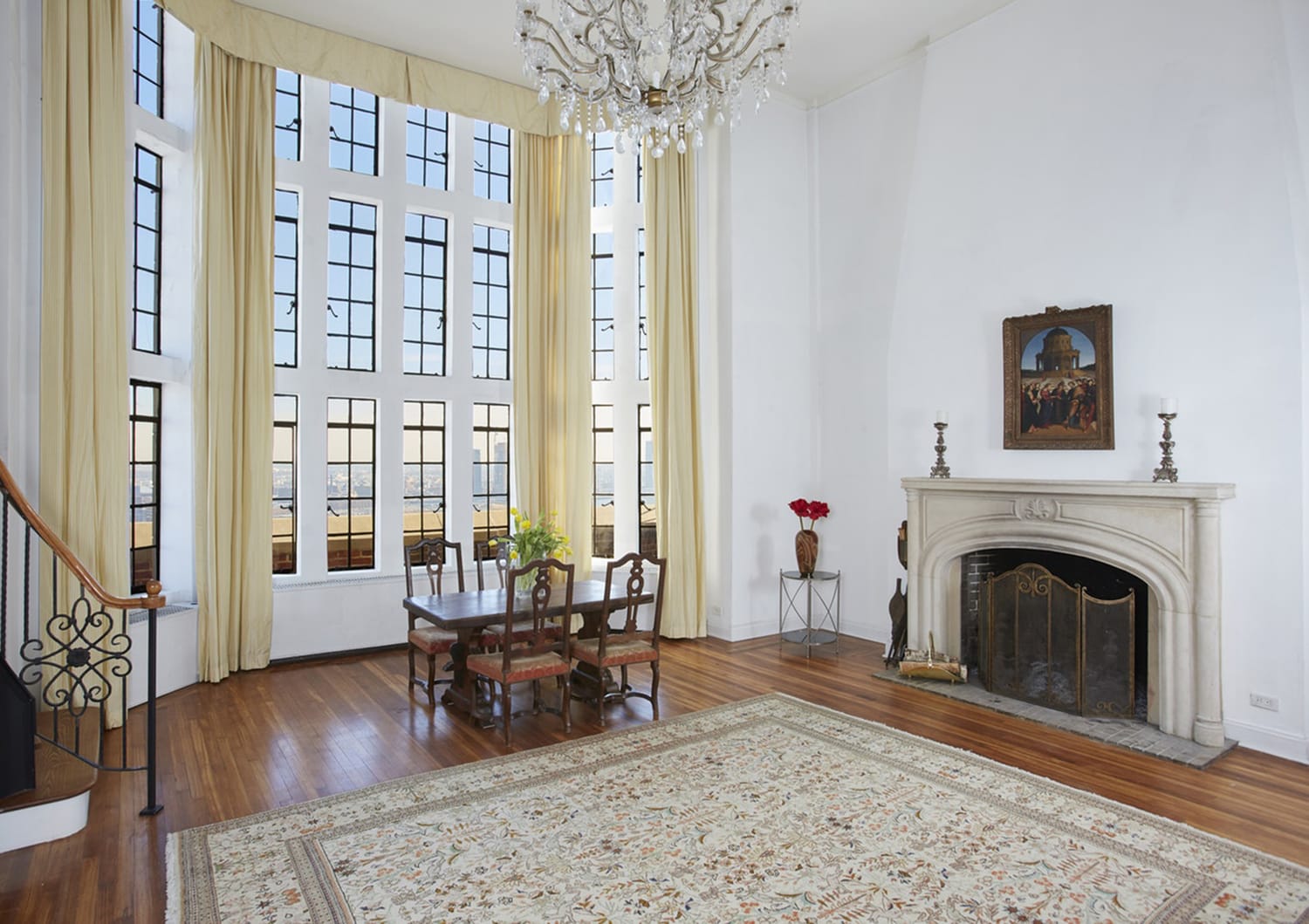 Spacious living room with large windows, elegant chandelier, dining table, ornate fireplace, and patterned rug.
