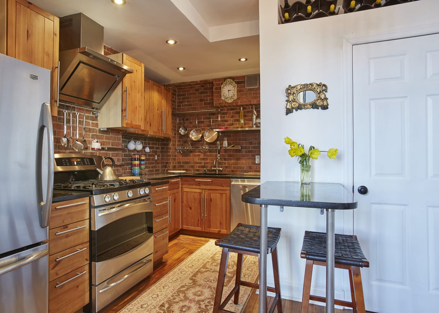 Modern kitchen with wooden cabinets, stainless steel appliances, small dining table, and vase with yellow flowers on top.
