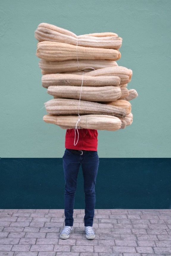 Person carrying a tall stack of bundled sisal drying mats standing against a muted green wall on a cobbled street.