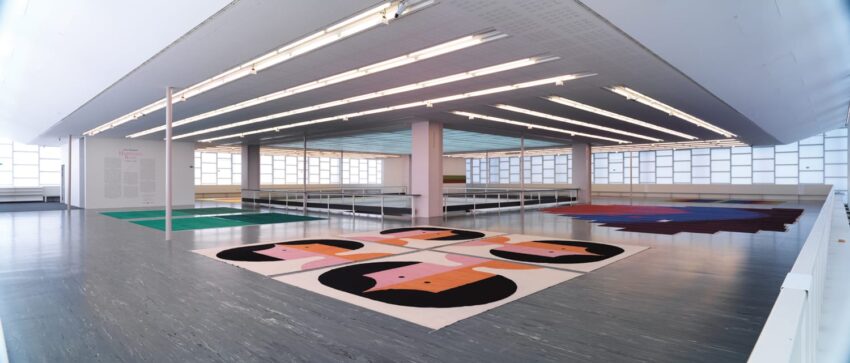 Modern art exhibition featuring colorful geometric rugs on a gallery floor with high ceilings and ample natural light.