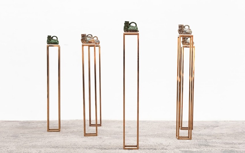 Ceramic sculptures on tall, narrow gold stands in a minimalist gallery setting with a concrete floor and white walls.