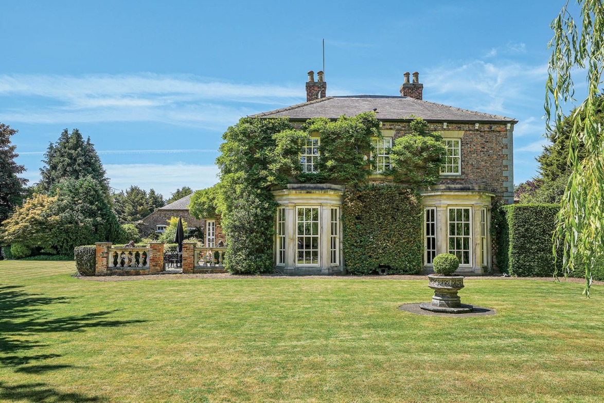 Stately brick house with ivy-covered walls, large windows, and manicured lawn under a clear blue sky.
