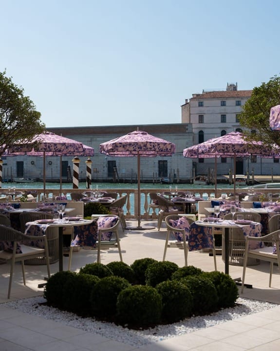 Outdoor restaurant with purple umbrellas and floral tablecloths overlooking a canal and buildings in the background.