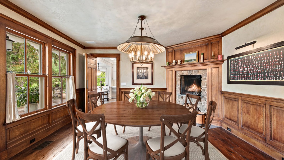 Elegant dining room with round wooden table, chairs, chandelier, fireplace, and large windows with garden view.