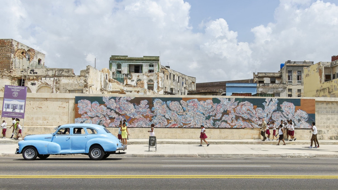 Vintage blue car on a street in front of a mural and old buildings, with pedestrians walking along the sidewalk under a cloudy sky.