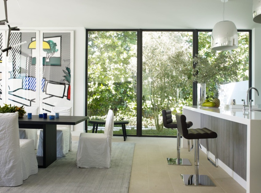 Modern dining area with large windows, contemporary art, and sleek furniture, overlooking a lush garden outside.