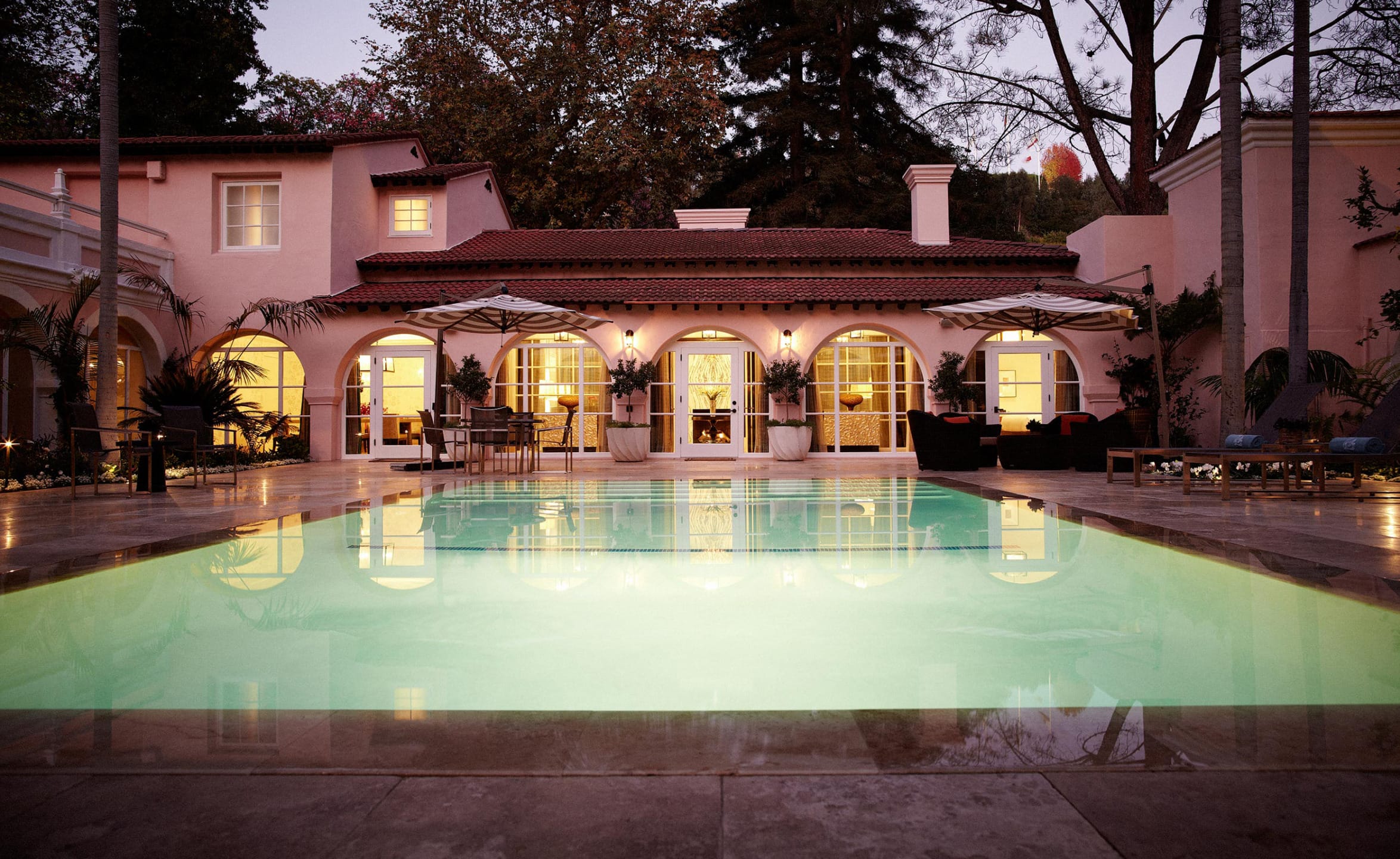 Luxurious villa with a lit swimming pool, arched windows, and palm trees at dusk.
