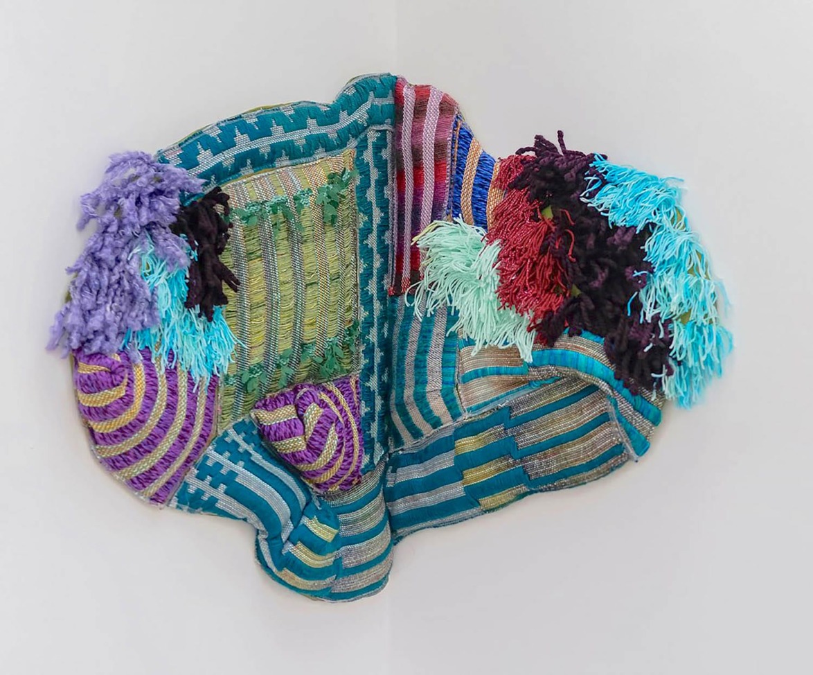 Colorful abstract textile art with various patterns and textures, featuring fringe and woven elements on a neutral background.