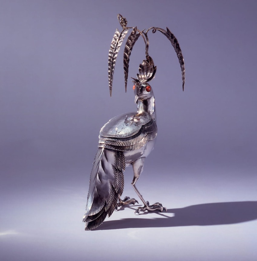 Silver bird sculpture with red eyes beneath a tall palm tree on a light purple background.