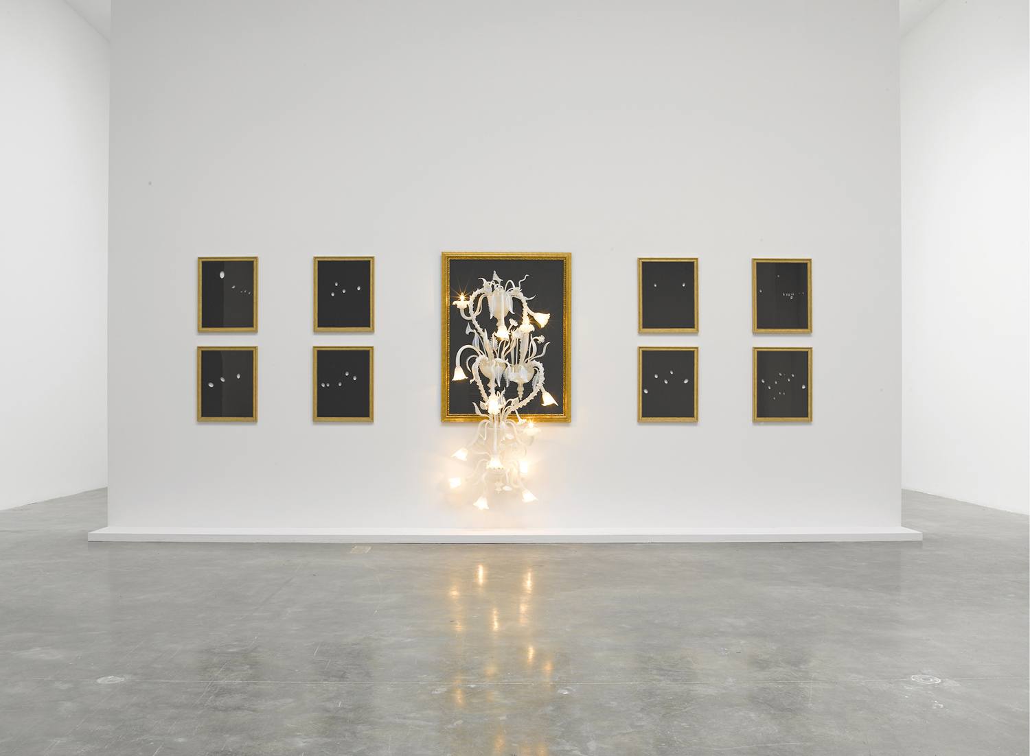 Minimalist art exhibition with nine framed abstract images and a lit sculptural piece displayed in a modern gallery setting.