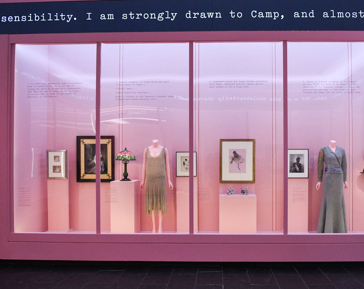 Museum exhibit display featuring framed art, vintage dresses, and accessories on mannequins against a pink backdrop.