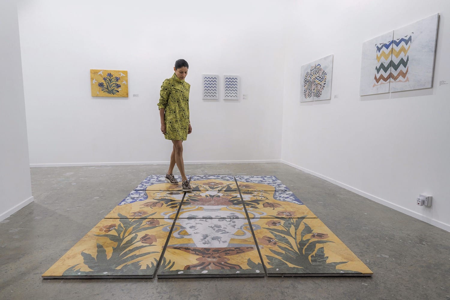 Woman walking on large patterned floor tiles in an art gallery with colorful artwork on white walls.