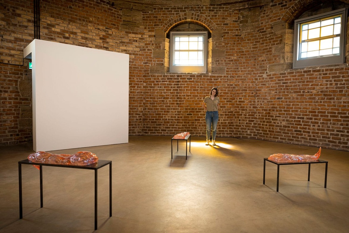 Art installation in a brick-walled room with three sculptures on tables and a person standing near a bright window.