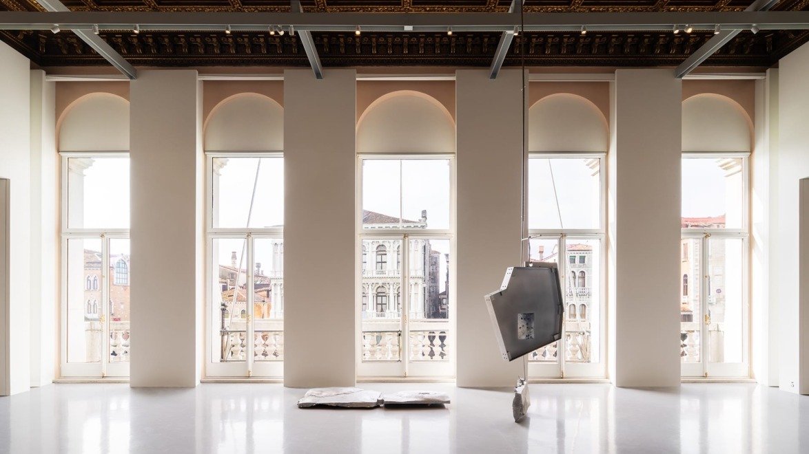 Spacious gallery with large windows, ornate ceiling, and contemporary metal sculptures on a reflective floor.