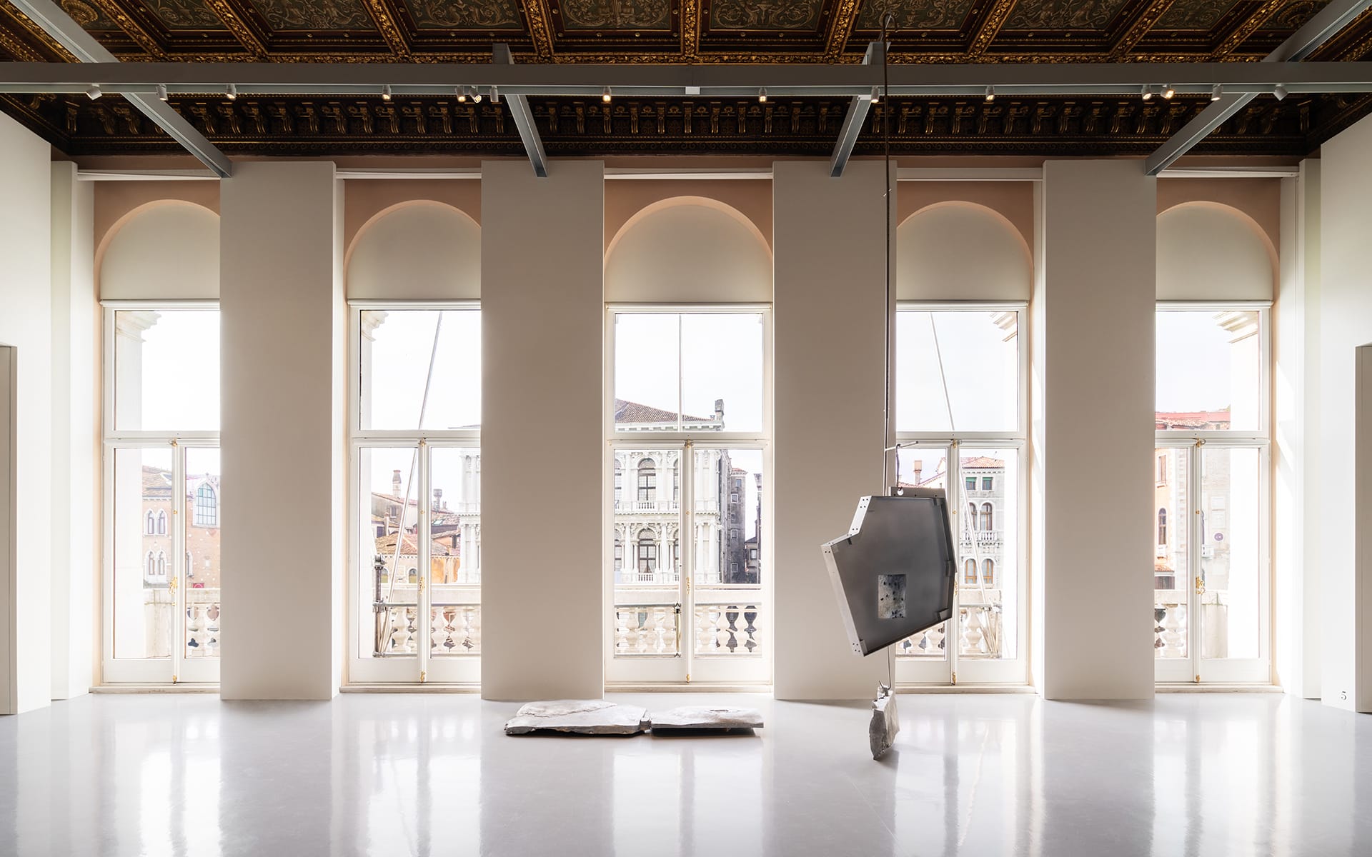 Spacious gallery with large windows, ornate ceiling, and contemporary metal sculptures on a reflective floor.