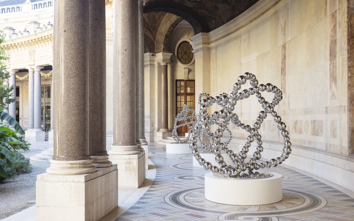 Large metal sculpture displayed on elegant stone walkway with columns and ornate architectural details in the background