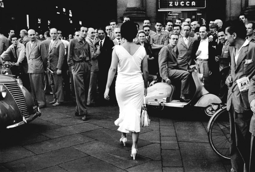 Woman in white dress walks past a crowd of men on a city street with vintage cars and scooters in the background.