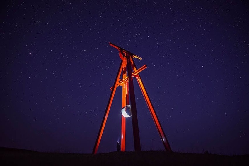 Large red sculpture under a starry night sky with a crescent moon, silhouetted figure at its base.