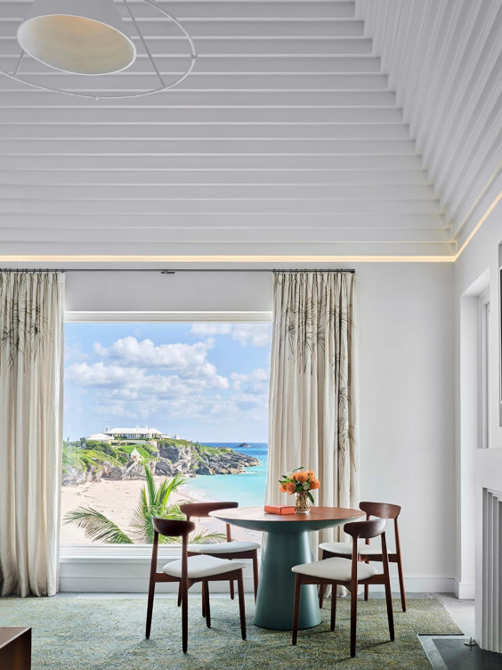 Modern dining area with round table, four chairs, and large window revealing scenic ocean and cliff view.