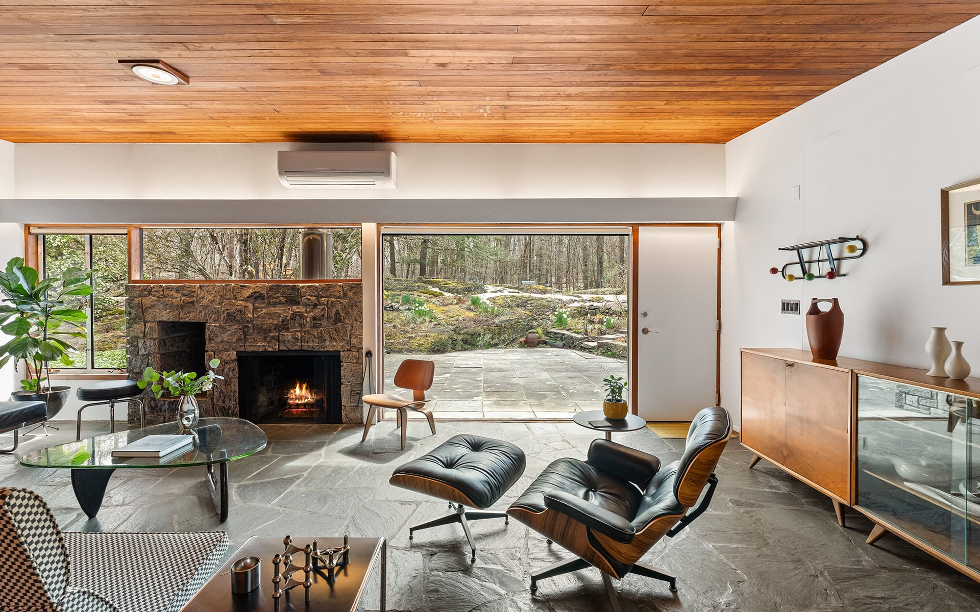 Mid-century modern living room with stone fireplace, sleek furniture, and large windows offering a forest view.