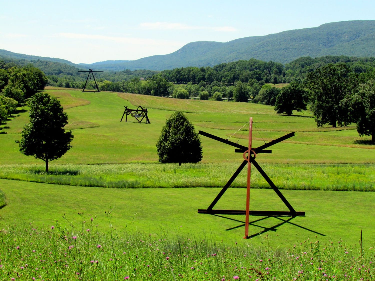 Large metal sculptures on grassy hills with trees and mountains in the background, under a clear blue sky.