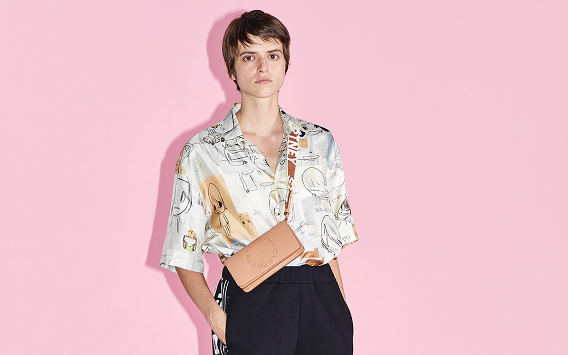 Person wearing a patterned shirt and crossbody bag against a pink background.