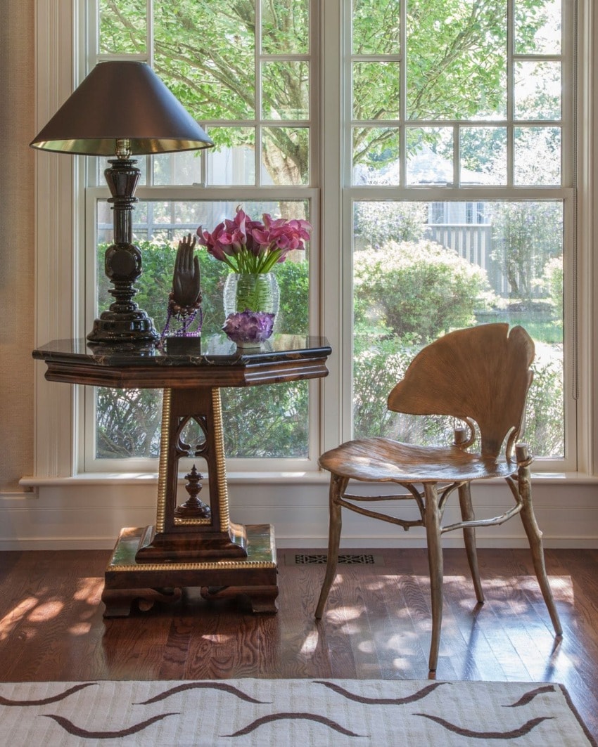 Sunlit room with wooden chair, elegant side table, lamp, and vase of pink tulips next to a large window with garden view.