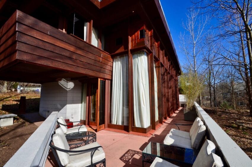 Exterior view of a modern house with wooden paneling, large windows, and a patio with white cushioned seating.