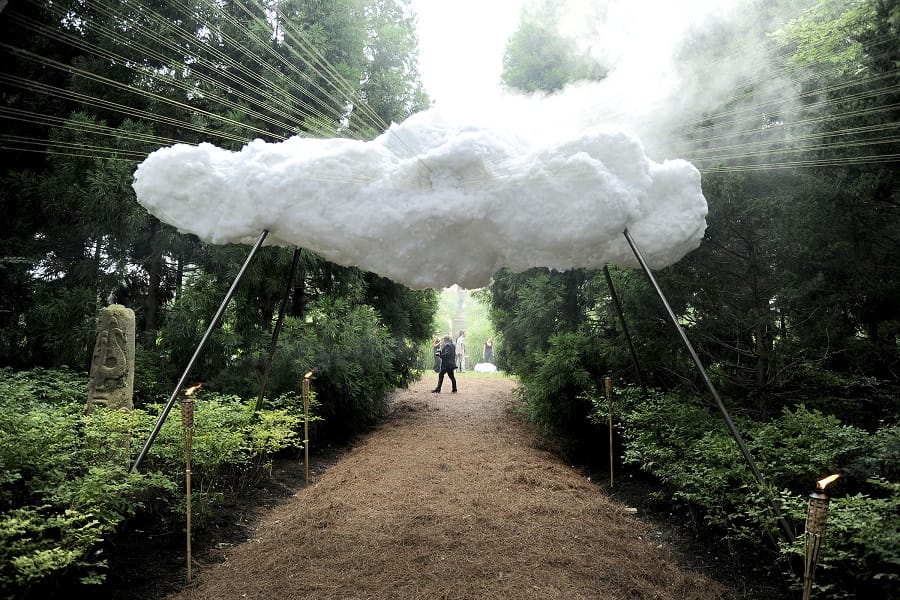 Artificial cloud installation suspended in a forest path, with steam creating a misty effect, surrounded by lush greenery.