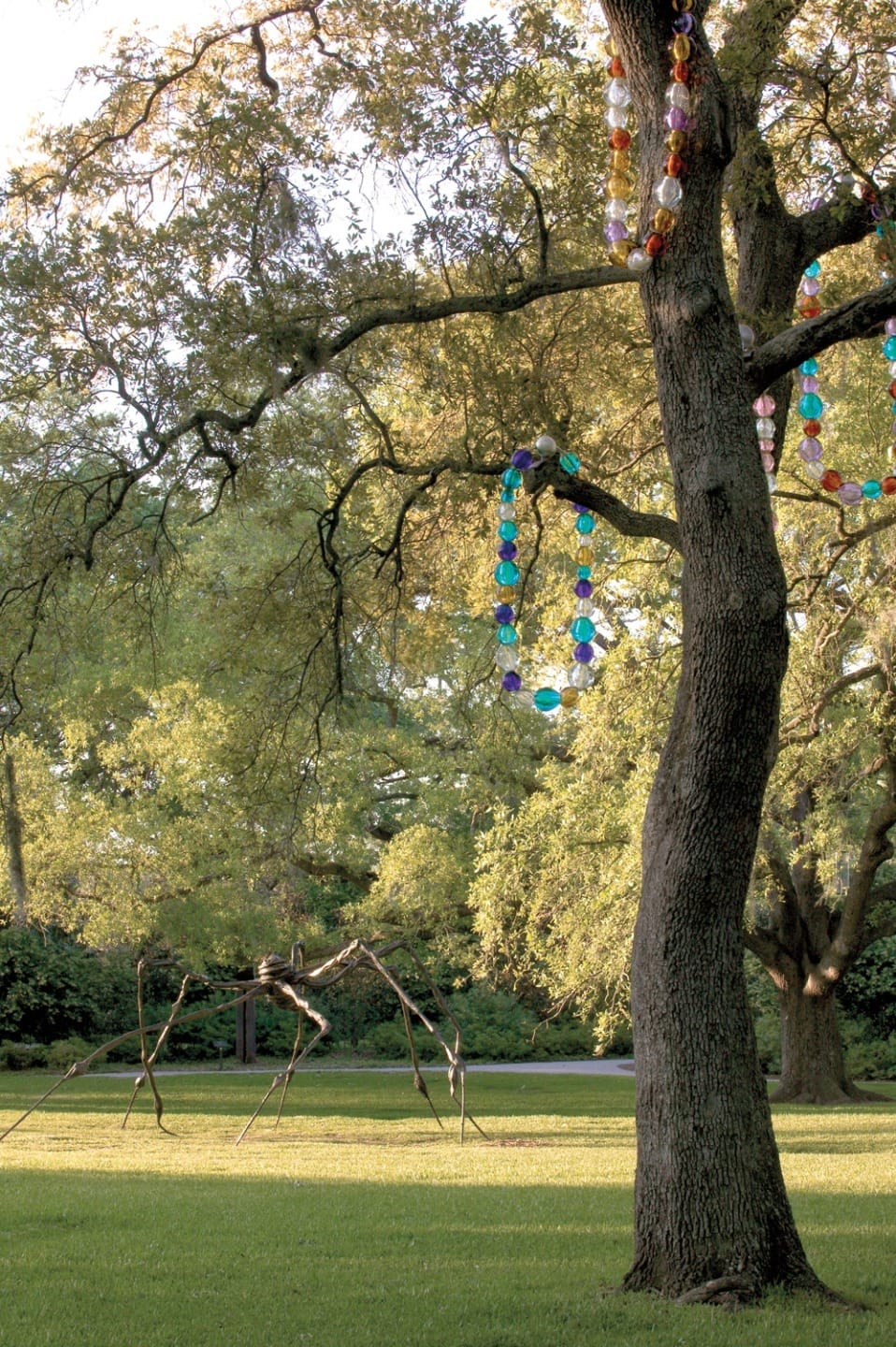 Tree decorated with colorful beads in a park with a sculpture resembling a spider in the background.